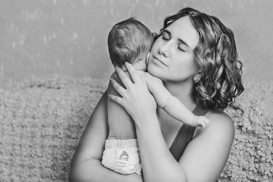 A mother with curly hair gently holds her little baby in her arms . Lifestyle at home with natural light.Black and white photo.