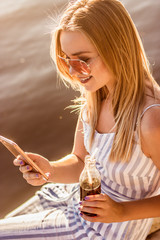 Portrait of happy young woman in pink sunglasses and stripped overalls uses mobile phone and drinks soda on beach. Beautiful blonde girl holds bottle of lemonade and smart phone in hands at riverside