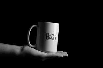 A mug that says "Super Daddy". A mug in his hands. Black background. Black picture.