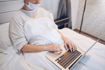 Unrecognizable young woman in face medicine mask  working on a laptop in bedroom during coronavirus isolation home quarantine. Covid-19 pandemic Corona virus. Distance online work from home concept.