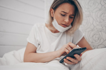 Woman in face protective mask lying in bed during coronavirus isolation home quarantine cleaning phone by hand sanitizer, using cotton wool with alcohol to wipe to avoid contaminating with COVID-19