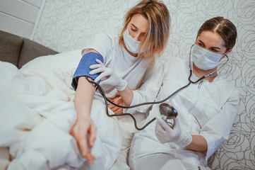Doctor nurse in protective face mask measures blood pressure with a sphygmomanometer. Coronavirus (COVID-19) concept. First symptoms. Woman sick of flu viral infection in home isolation quarantine