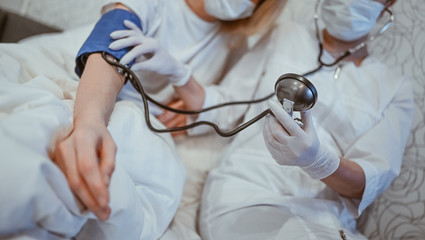 Doctor nurse in protective face mask measures blood pressure with a sphygmomanometer. Coronavirus (COVID-19) concept. First symptoms. Woman sick of flu viral infection in home isolation quarantine