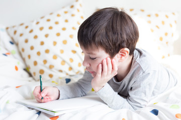 Naklejka premium School Boy on bed and doing school homework at home, writing in notebook. Distance learning online education. Quarantine