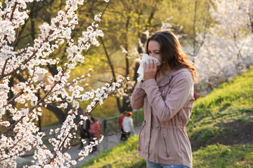 Young pretty woman sneezing in a park with blooming trees in spring. Pollen Allergy Symptoms