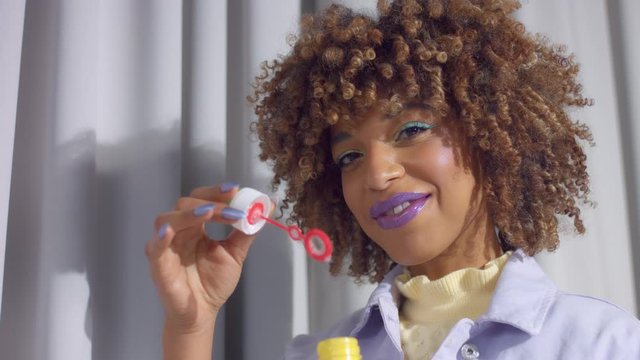 Mixed race woman on grey curtains background in pastel colors clothes dancing and blows to a bubble blower making flying a soap bubbles