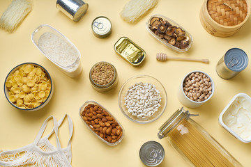 Creative flatlay with pantry staples. Glass jars with pasta, beans and chickpeas, canned goods, nuts and dried mushrooms in reusable containers. Top view pattern with basic products 