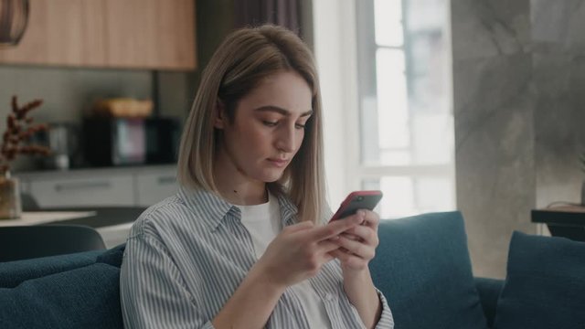 Serious pretty young european woman in white t-shirt sit on sofa at modern room looking at cellphone screen working reading or watching news chatting in social media surfs the inernet slow motion