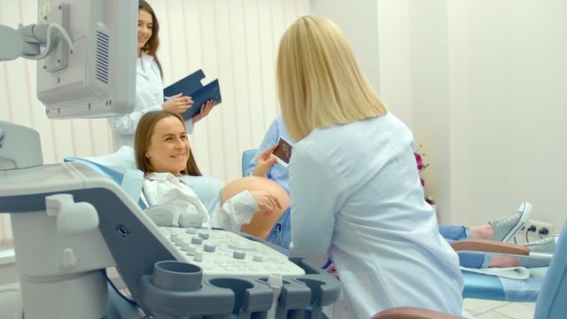 Beautiful pregnant woman lying on examination coach, sharing ultrasound images of baby with husband while female doctor giving consultation. Video in 4K