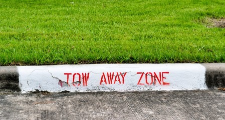 Tow Away Zone sign painted in red letters on a curb at a through way with green trimmed lawn behind it.