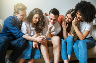 Group of friends sitting on the sofa at home watching a woman's smart phone - Millennials have fun together in an apartment