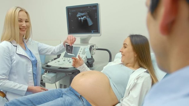 Beautiful pregnant woman lying on examination coach, sharing ultrasound images of baby with husband while female doctor giving consultation. Video in 4K