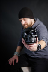 young adult man with a beard in a black hat holds a black skull in his hands