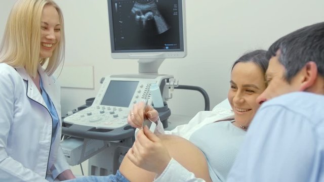 A woman doctor gives photos of an ultrasound scan to a pregnant woman. The girl was expecting a newborn baby. Happy couple watching a picture and smiling