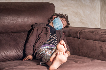 Latino boy in striped pajamas sick with covid-19, sitting on a brown sofa wearing a respirator, with dark hair and head on one side, long dark hair and feet without socks.