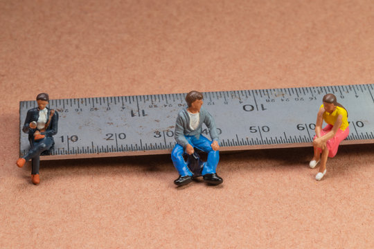 Miniature figures sit on a metal rule or ruler demonstrating the concept of social distancing to avoid transmission of the corona virus or other contagious illness