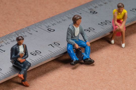 Miniature figures sit on a metal rule or ruler demonstrating the concept of social distancing to avoid transmission of the corona virus or other contagious illness
