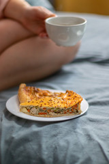 Tasty vegetable piece of pie on a white glass plate on the bed. Girl with a cup of hot coffee sits on a gray bed. Breakfast in bed, cozy hotel room. Vertical photo