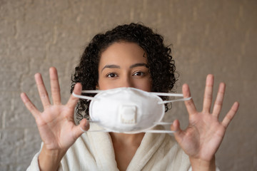 Brazilian black woman showing face mask to prevent to spread CoronaVirus. Girl at home wearing white robe and holding protective equipment. Global pandemic. Coronavirus quarantine.