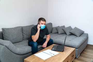 Quarantine coronavirus COVID19 distance job. Young businessman in mask talking on smart phone, sitting with laptop. Office man freelancer having conversation on cell phone while working from home