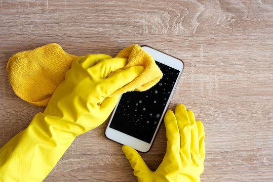 Female hand in rubber protective glove with rag wiping smartphone using cleaning spray.
