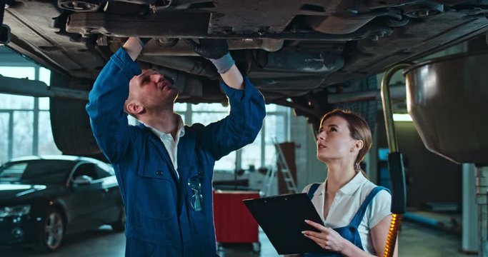 Man teaching young woman to repair car breaks. Male doing technical review in auto service from under and teaching female colleague. Woman learning and noting or filling in document in garage.