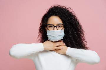Sick curly haired woman touches neck, suffers from suffocation and shortage of breathing, wears medical mask to avoid virus infection, isolated on pink background. Coronavirus, health care concept