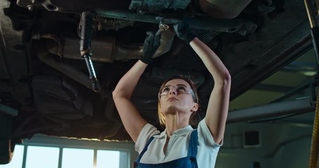 Caucasian young beautiful woman in uniform and goggles standing under car and twisting some detail with wrench in garage. Female mechanic working in auto service salon and repair breaks.