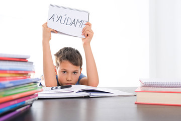 Tired frustrated boy sitting at the table with many books, exercises books. Spanish word Auydame -...