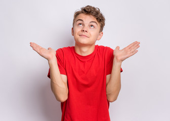 Teen boy showing helpless gesture with hands - I do not know. Cute young teenager in red t-shirt, on grey background. Shrugging, confused child making helpless sign and looking up.