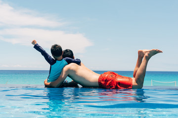Rear view of father and son looking at sea while sitting by swimming pool against sky