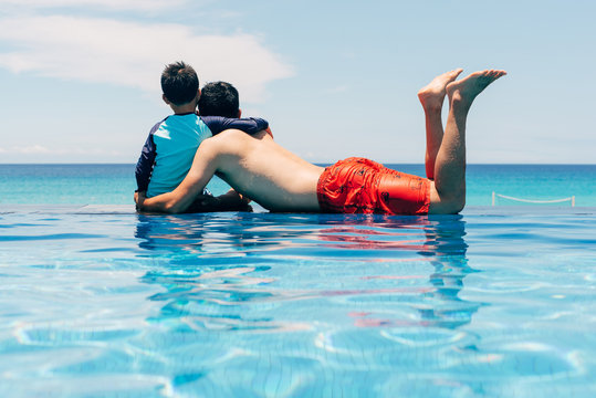 Rear view of father and son looking at sea while sitting by swimming pool against sky