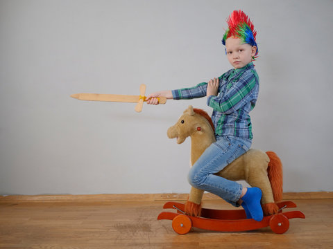 happy boy in plaid shirt is rolling on rocking horse toy. baby with wonderful punk-style colored hairdo. kid is armed with wooden sword and brandishes it. Dreams about travel and heroic exploits