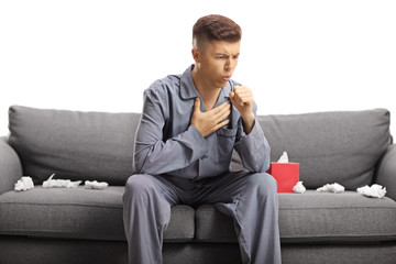 Young man coughing and sitting on a sofa in pajamas with tissue papers