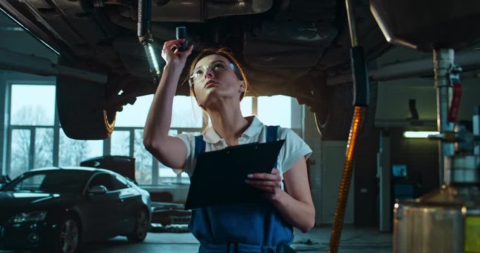 Caucasian woman standing under car and checking it with flashlight in garage. Female mechanic working in auto service and doing technical review of automobile. Worker noting and filling in document.