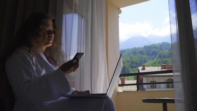 Woman silhouette with long hair in white bathrobe sitting near window. Pretty girl working on laptop and using smartphone in hotel apartment enjoying amazing landscape outside room window closeup