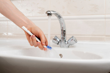 a close up hand holding a white toothbrush and wash it under the water in sink in bathroom