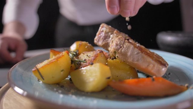 Cooks hand puts a fresh fried steak on white plate and maiking final decoration of dish. The cook sprinkles meat with salt, pepper, seasoning, spices and lemon juice. Serving food.