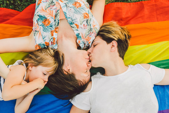 Lesbian couple kissing besides their little daughter in a summer day while lying over a lgbt flag