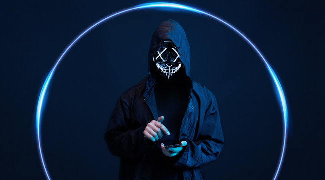 Dangerous hooded hacker in mask with smartphone in his hand. Internet, cyber crime, cyber attack, system breaking and malware concept. Anonymous. Dark background.