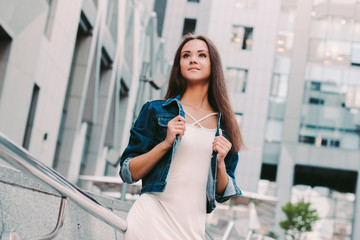 Portrait of cute beautiful stylish hipster girl posing outdoors among urban buildings. Attractive fashionable young woman in denim jacket and dress walking city streets. Modern female outfit for city