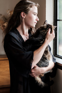 Young woman looking through window while patting cat at home