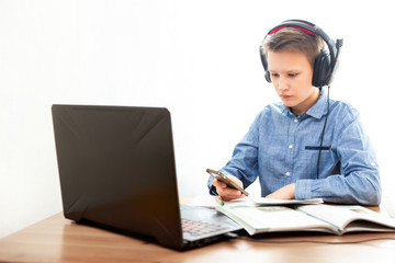 A boy in headphones in front of a computer does his homework. The boy is holding a phone. Distance learning in quarantine.