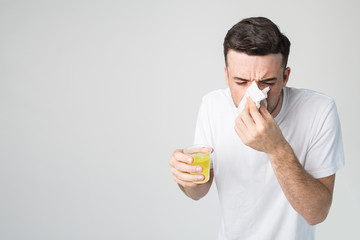 Young man isolated over background. Sick person with yellow liquid in glass sneezing into white napkin. Suffer from sickness or coronavirus danger. Alone in studio.