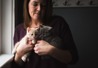 Woman holding two adorable kittens in her arms next to a window.