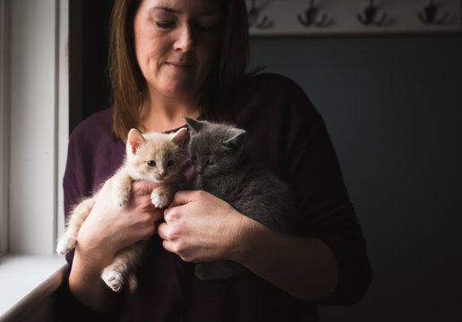 Woman holding two adorable kittens in her arms next to a window.