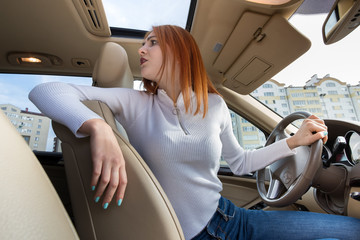 Wide angle view of young redhead woman driver driving a car backwards looking behind.