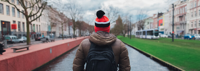 Perspective panoramic banner, back view of young guy, tourist, wearing colorful hat and backpack on background of city river.