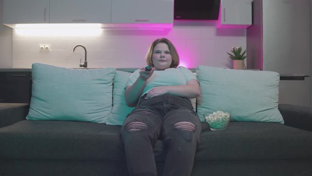 Smiling plump teenager changing tv channels by remote control, weekend leisure