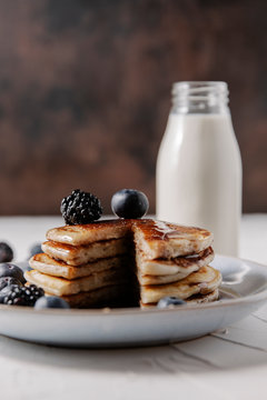 Still life of a stack of homemade pancakes with berries and syrup on a plate, and a bottle glass of milk in background.